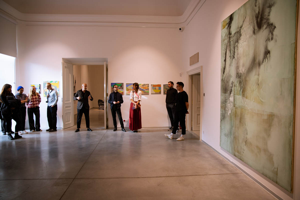 Ludzie w przestrzeni wystawowej, naścianie są prezentowane abstrakcyjne grafiki