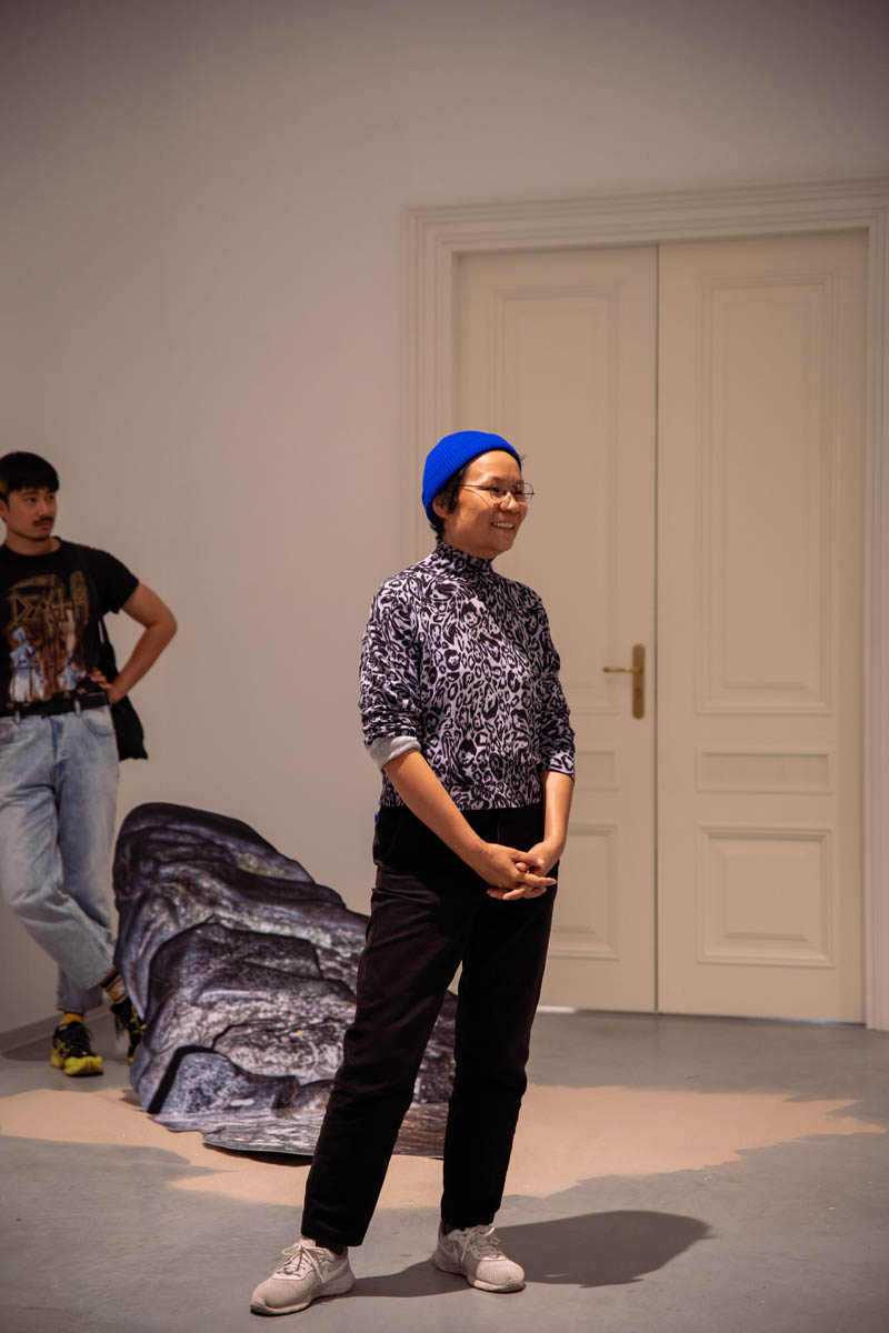 Uśmiechnięta uczestniczka wernisażu stoi w przestrzeni wystawowej. Za nią na podłodze jedna z prezentowanych prac.