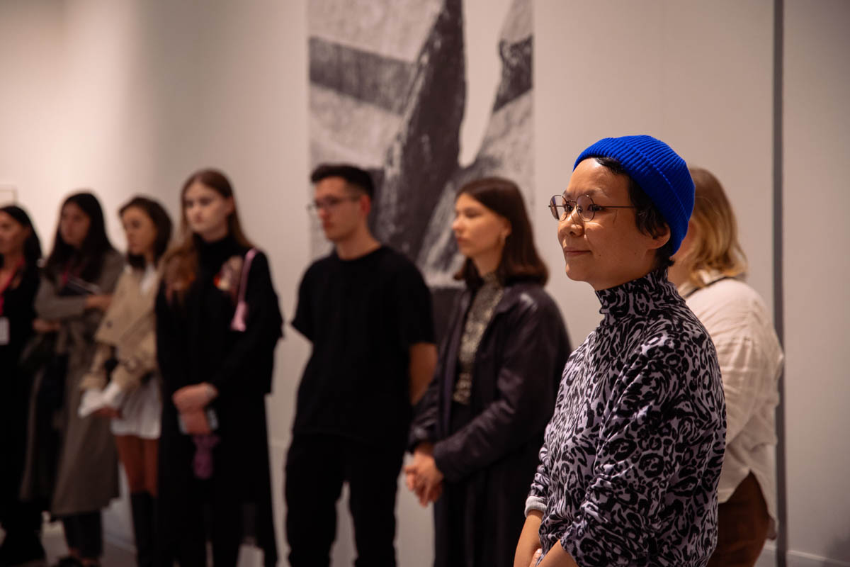 Zbliżenie na uczestniczkę wernisażu, w tle stoją artyści, których prace są prezentowane na wystawie