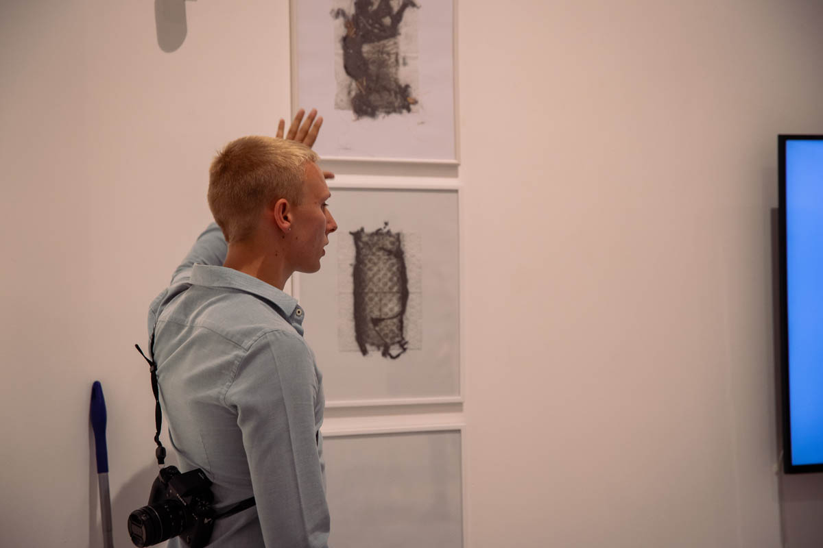 Gość wernisażu z uniesioną dłonią opisuje abstrakcyjne grafiki