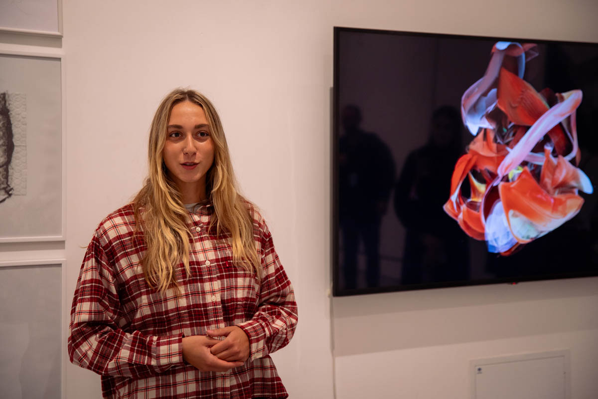 Uczestniczka wernisażu, za nią na dużym monitorze prezentowana na ciemnym tle wielobarwna abstrakcyjna grafika