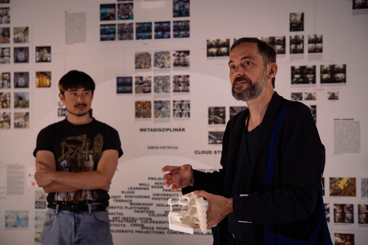 Mężczyzna opisuje prezentowane za nim geometrycznie zawieszone na ścianie zdjęcia, za nim ze skrzyżowanymi rękami stoi chłopak