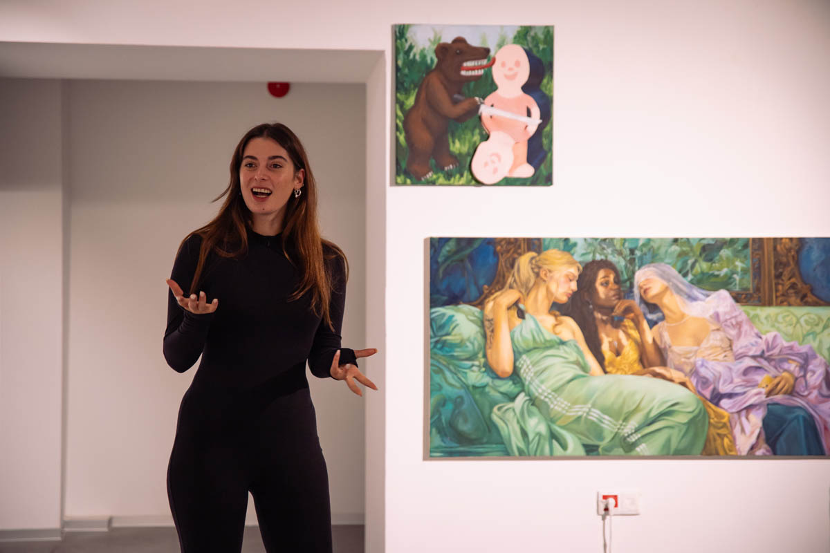 Uśmiechnięta młoda kobieta uchwycona w ruchu z uniesionymi dłońmi opisuje prezentowane za nią obrazy