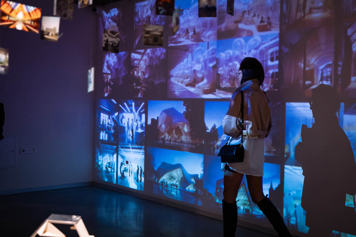 Uczestniczka wernisażu przechodzi wzdłuż ściany z wyświetlanymi z projektora zdjęciami. Obraz z projektora pada także na jej sylwetkę