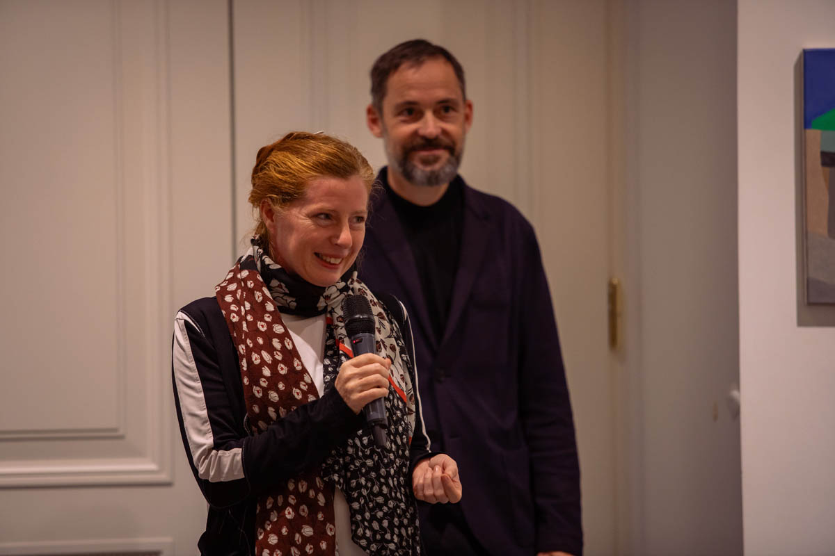 Otwarcie wernisażu. Przez mikrofon przemawia kobieta, za nią stoi uśmiechnięty mężczyzna