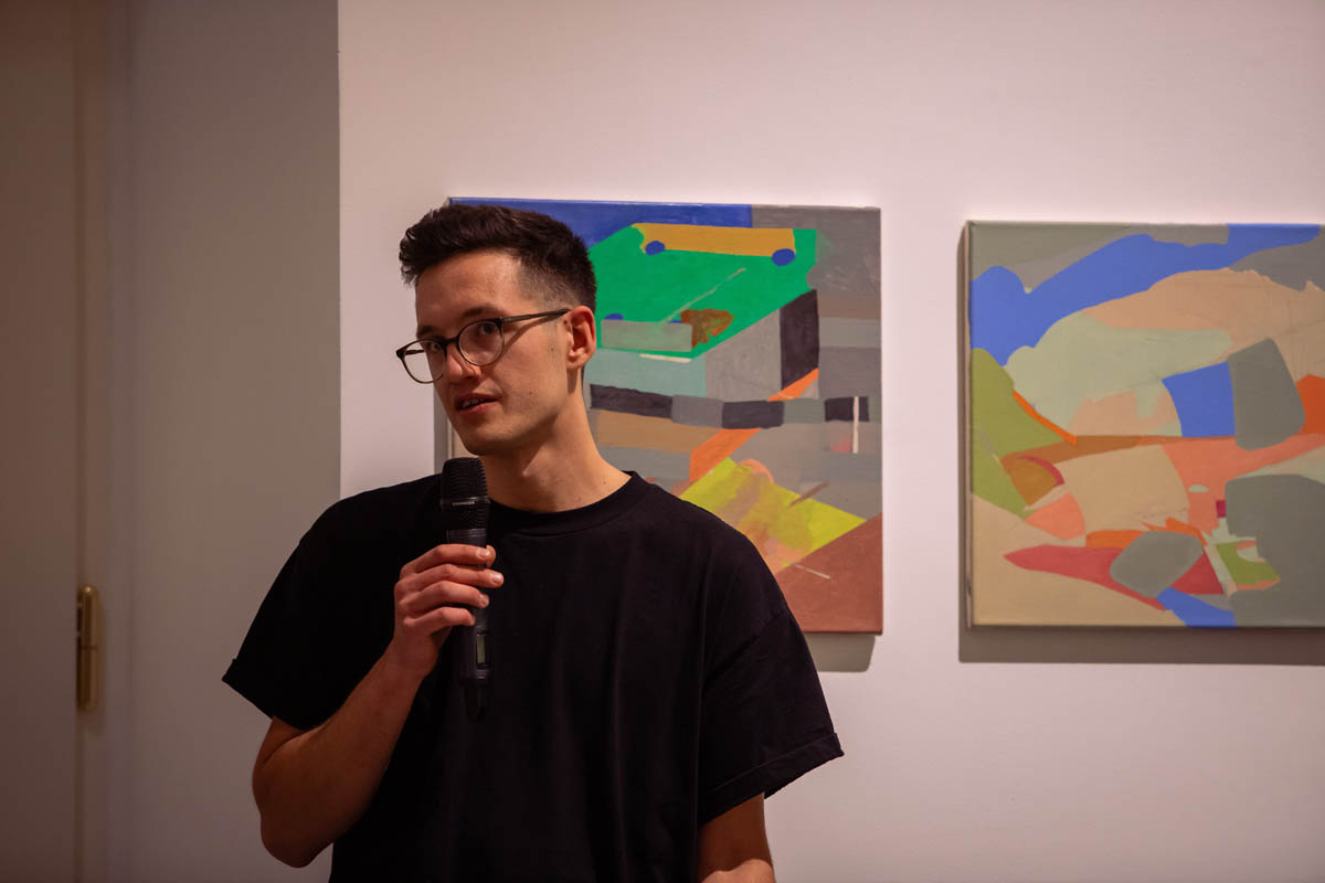 Młody mężczyzna przemawia przez mikrofon. Za nim na ścianie wielokolorowe geometryczne abstrakcyjne grafiki