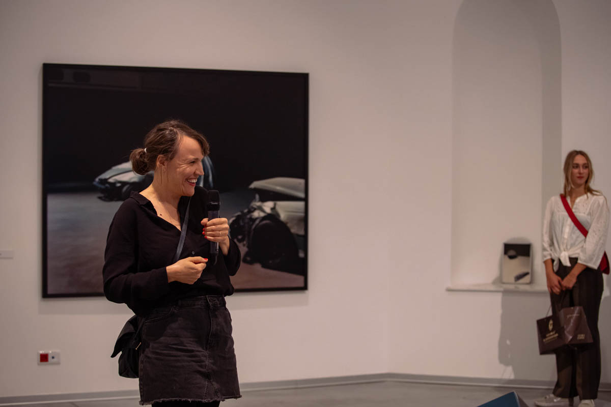 Uśmiechnięta kobieta z mikrofonem, w tle na ścianie jedna z prezentowanych prac