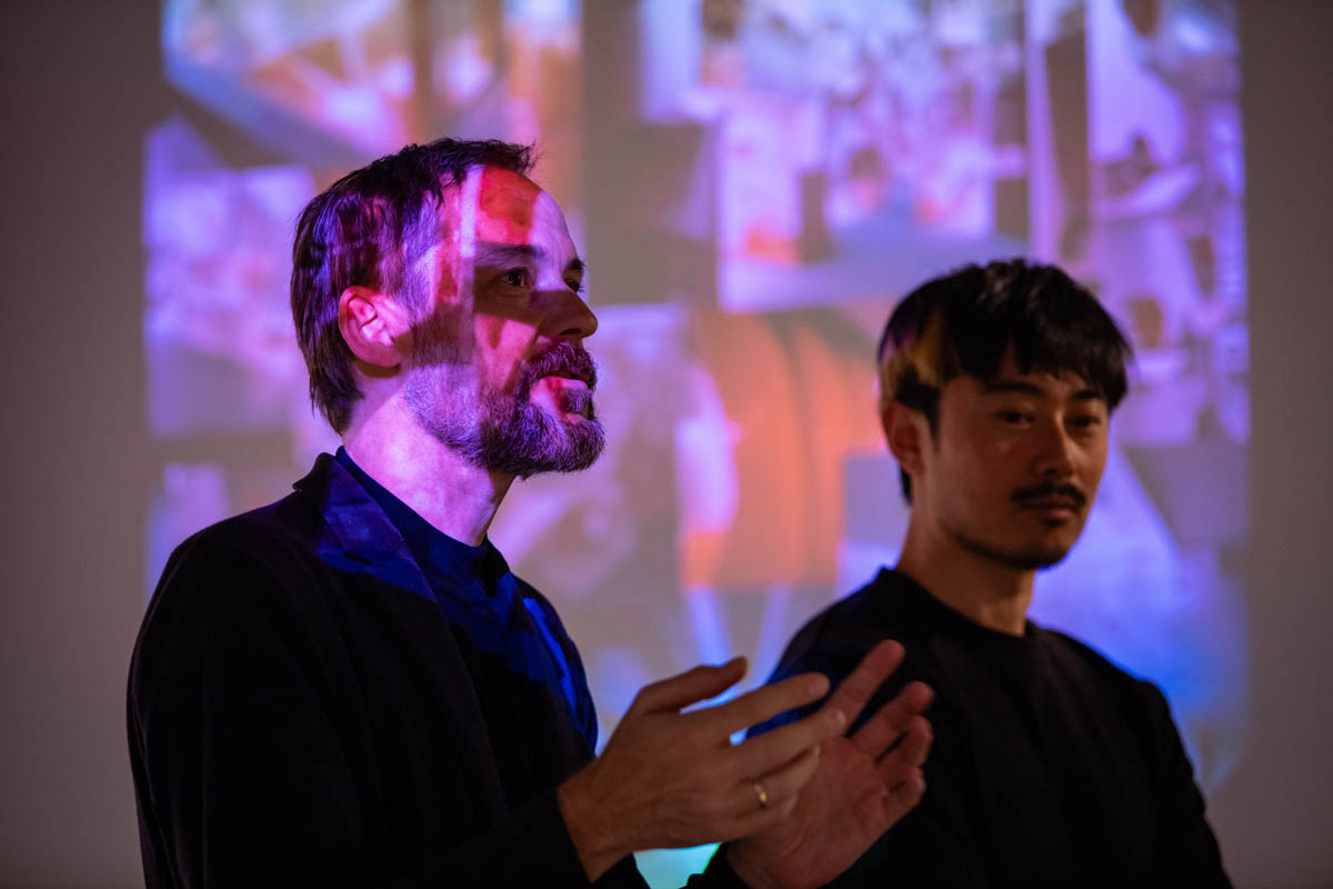 Dwóch mężczyzn w przestrzeni wystawowej. Starszy omawia pracę, jego twrz jest oświetlona kolorowym obrazem z projektora