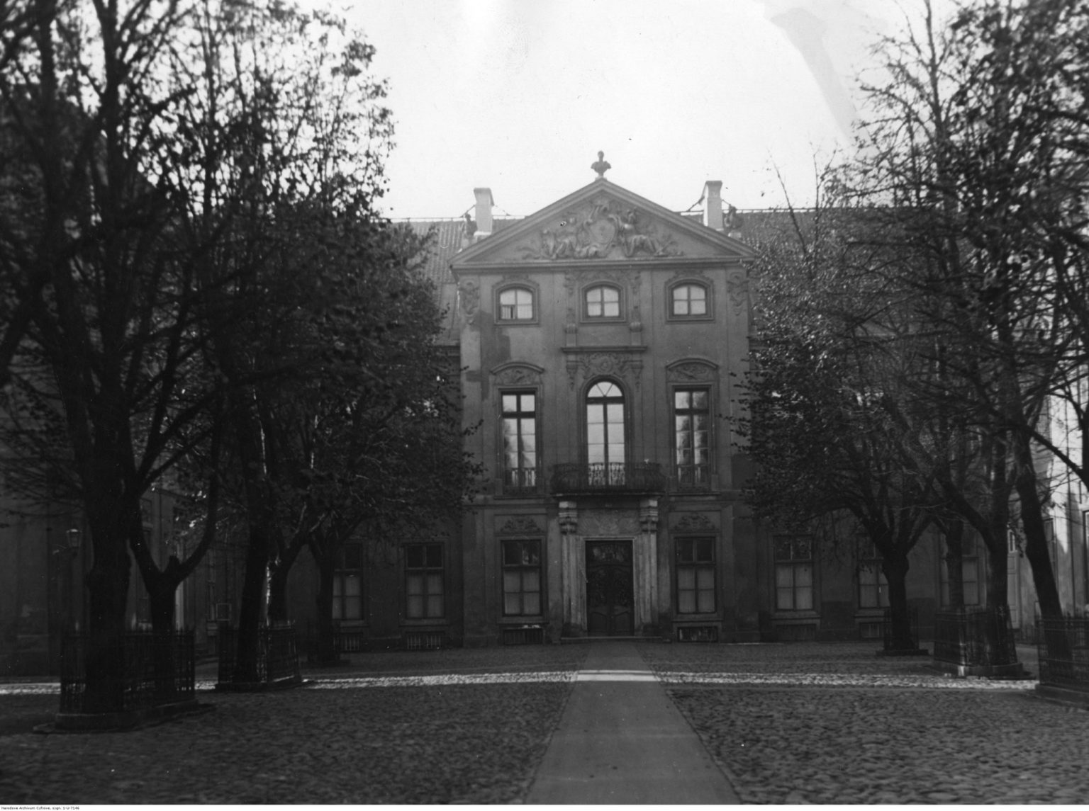 Widok fasady pałacu, przysłoniętej drzewami, rosnącymi po obu stronach alejki biegnącej przez środek dziedzińca