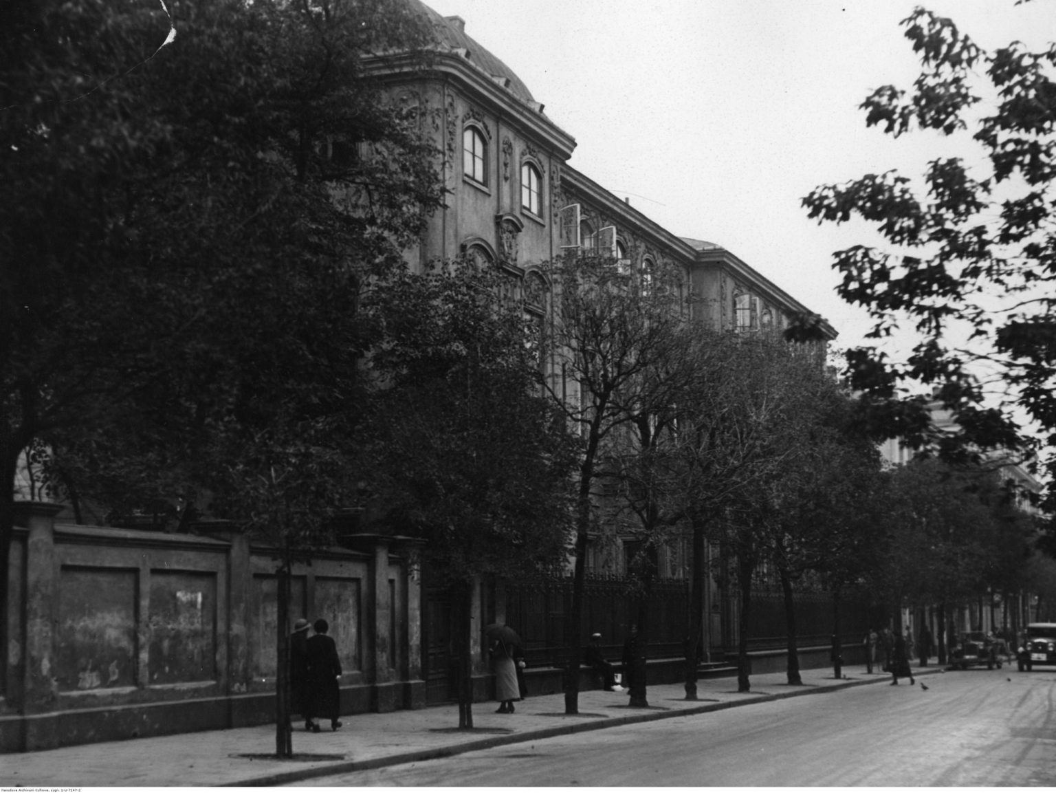 Widok budynku od strony ulicy. Budynek przesłonięty jest posadzonymi wzdłuż ulicy drzewami