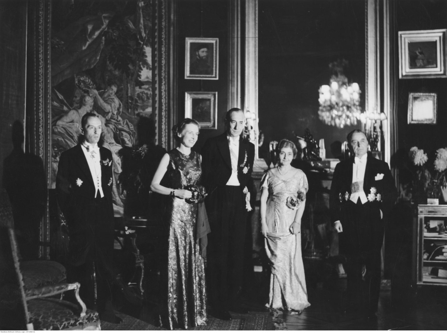 3 mężczyzn w garniturach i 2 kobiety w sukniach pozują do zdjęcia we wnętrzu pałącu. Na ścianie za nimi wiszą obrazy