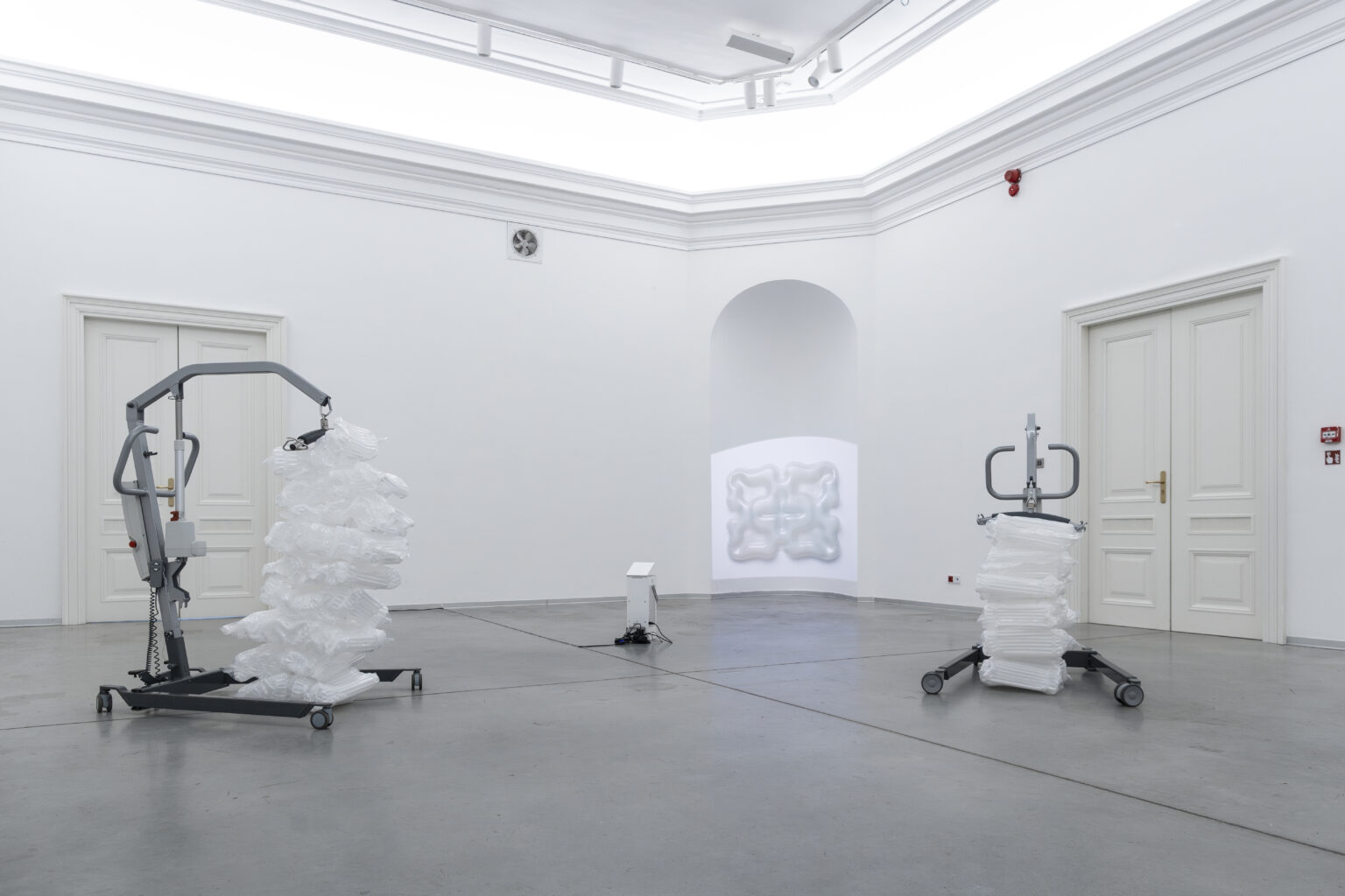 Po prawej i lewej stronie urządzenia do ćwiczeń, przytrzymujące białe, pompowane elementy wygenerowane przez sztuczną inteligencję. W tle obraz rzucany z projektora