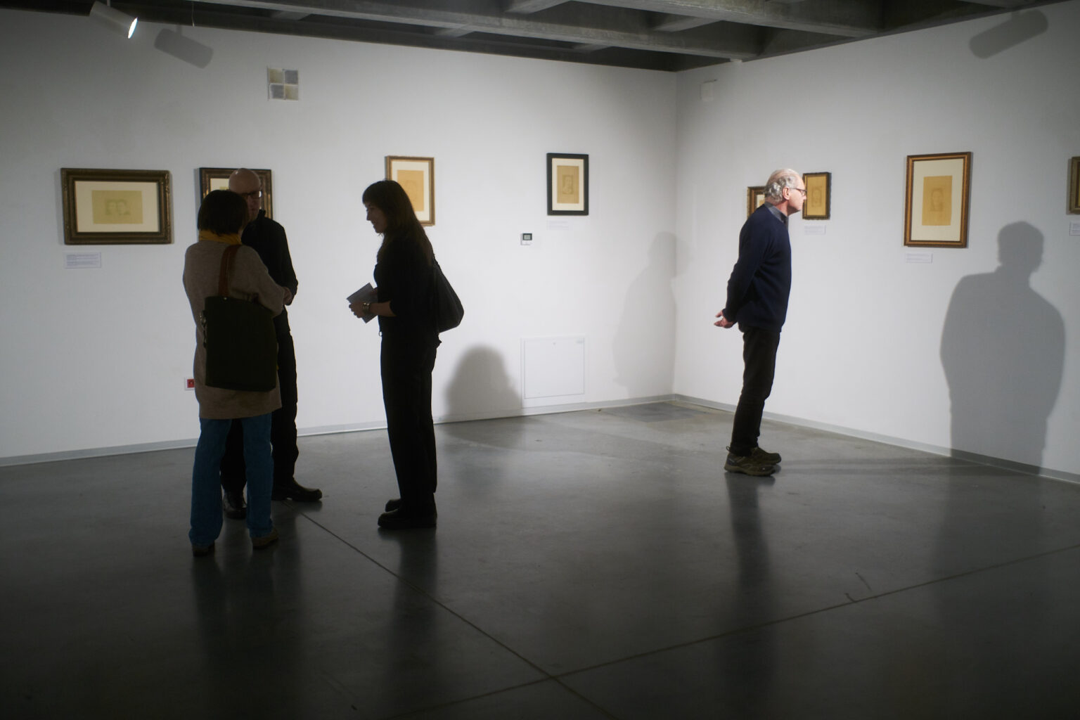 Słabo oświetlona sala w galerii sztuki. Na ścianach podświetlone oprawione w ramy grafiki. Przed nimi stoją ciemne figury ludzi