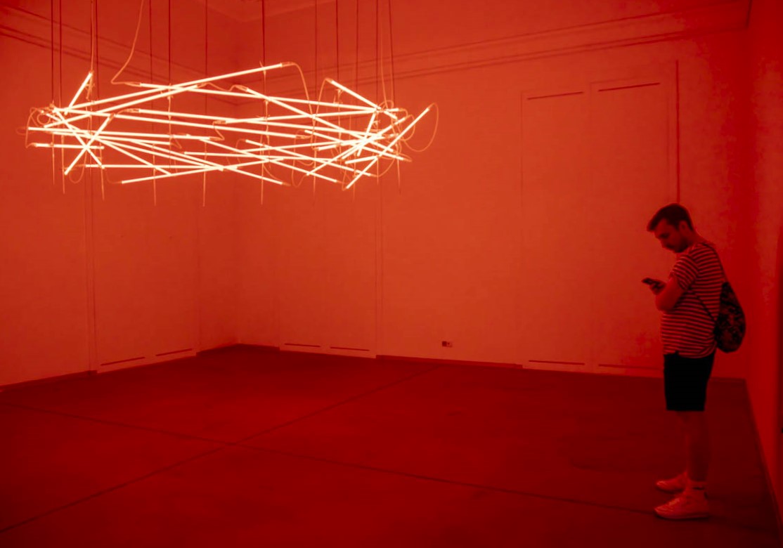 Mężczyzna stoi w oświetlonej na czerwono sali wystawowej. Z sufitu wiszą świecące splątane jarzeniówki