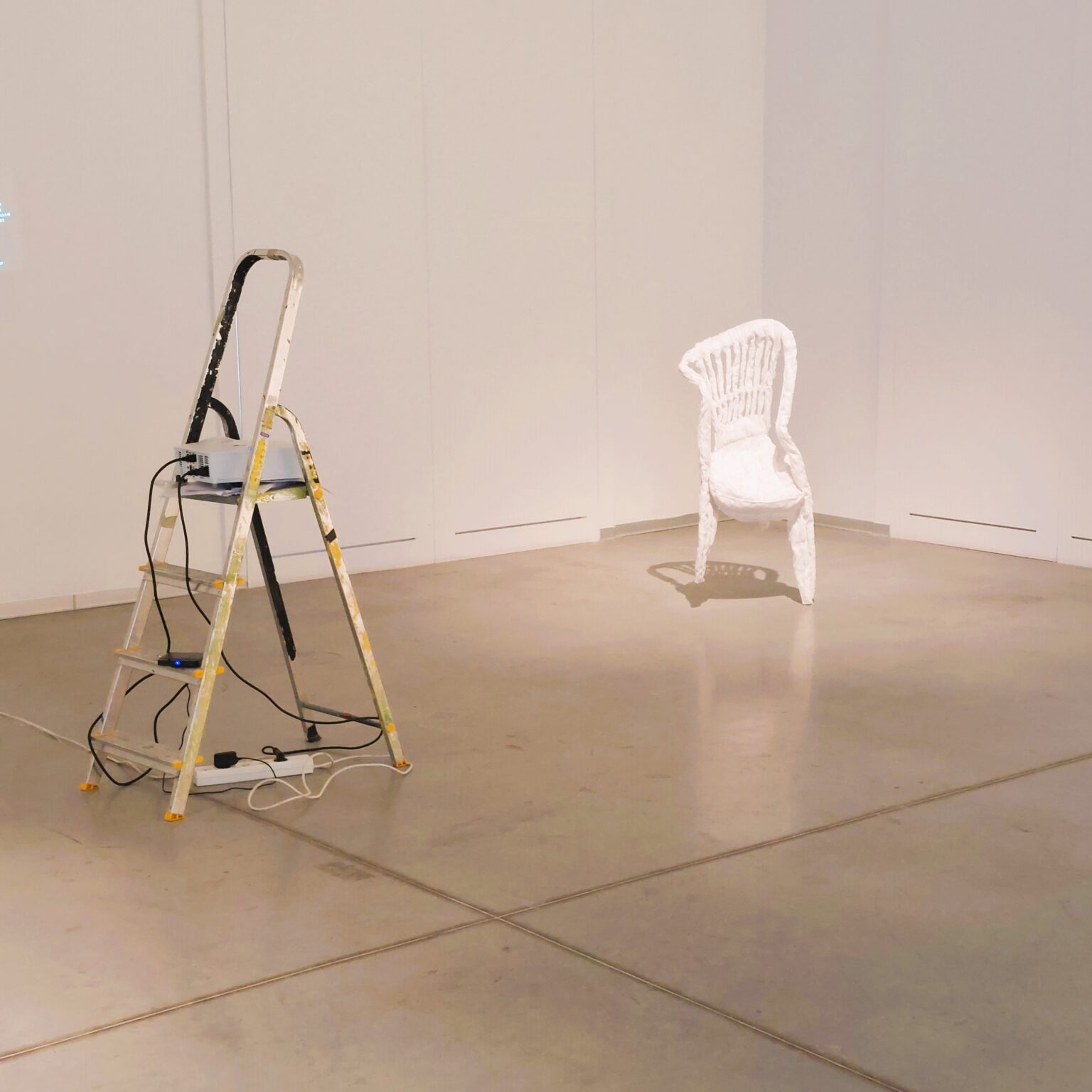 Białe krzesło w rogu sali. Na środku drabina z projektorem.