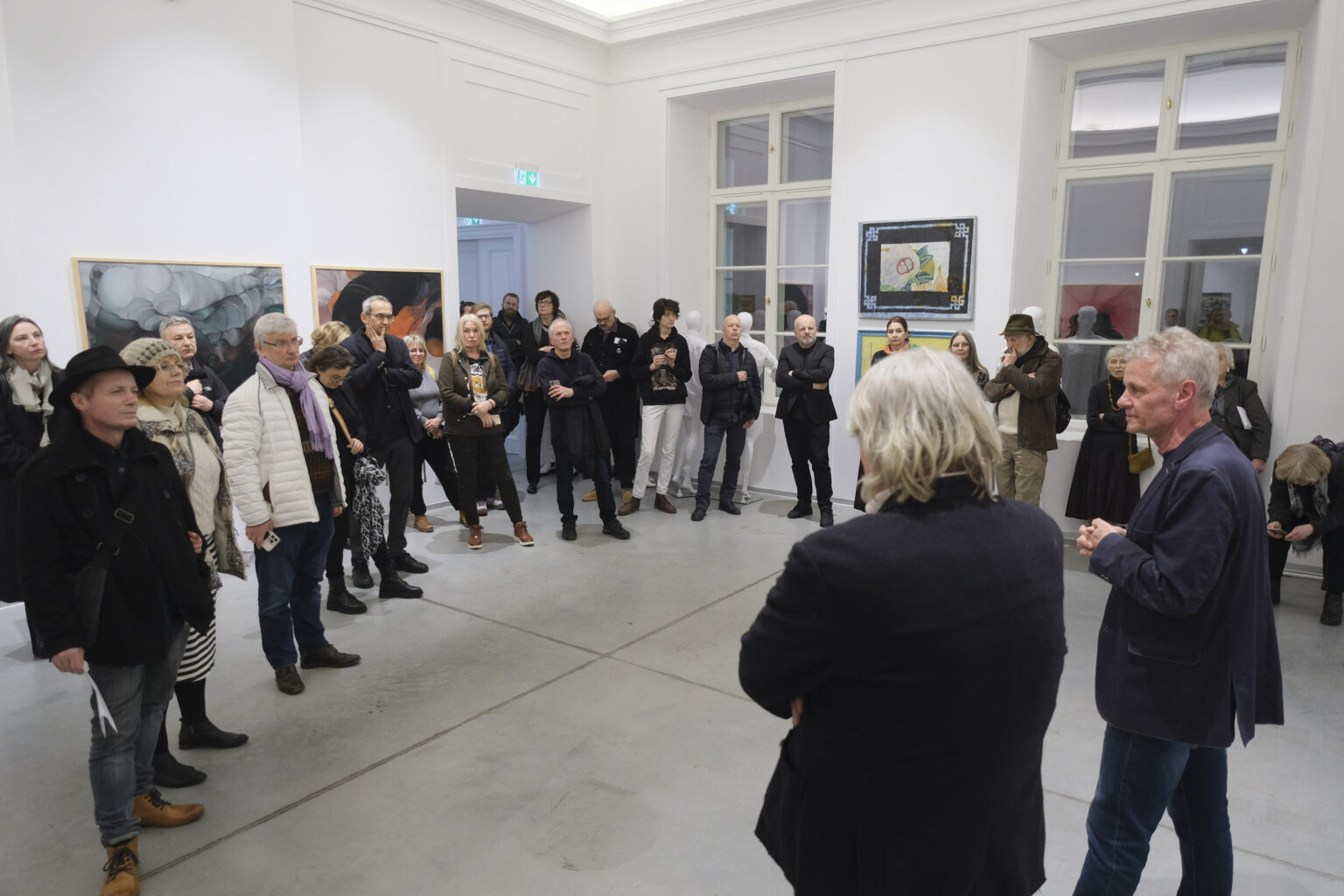 Widzowie wystawy stoją w większej grupie w przestrzeni galerii. Na ścianach wiszą obrazy Konrada Jarodzkiego.