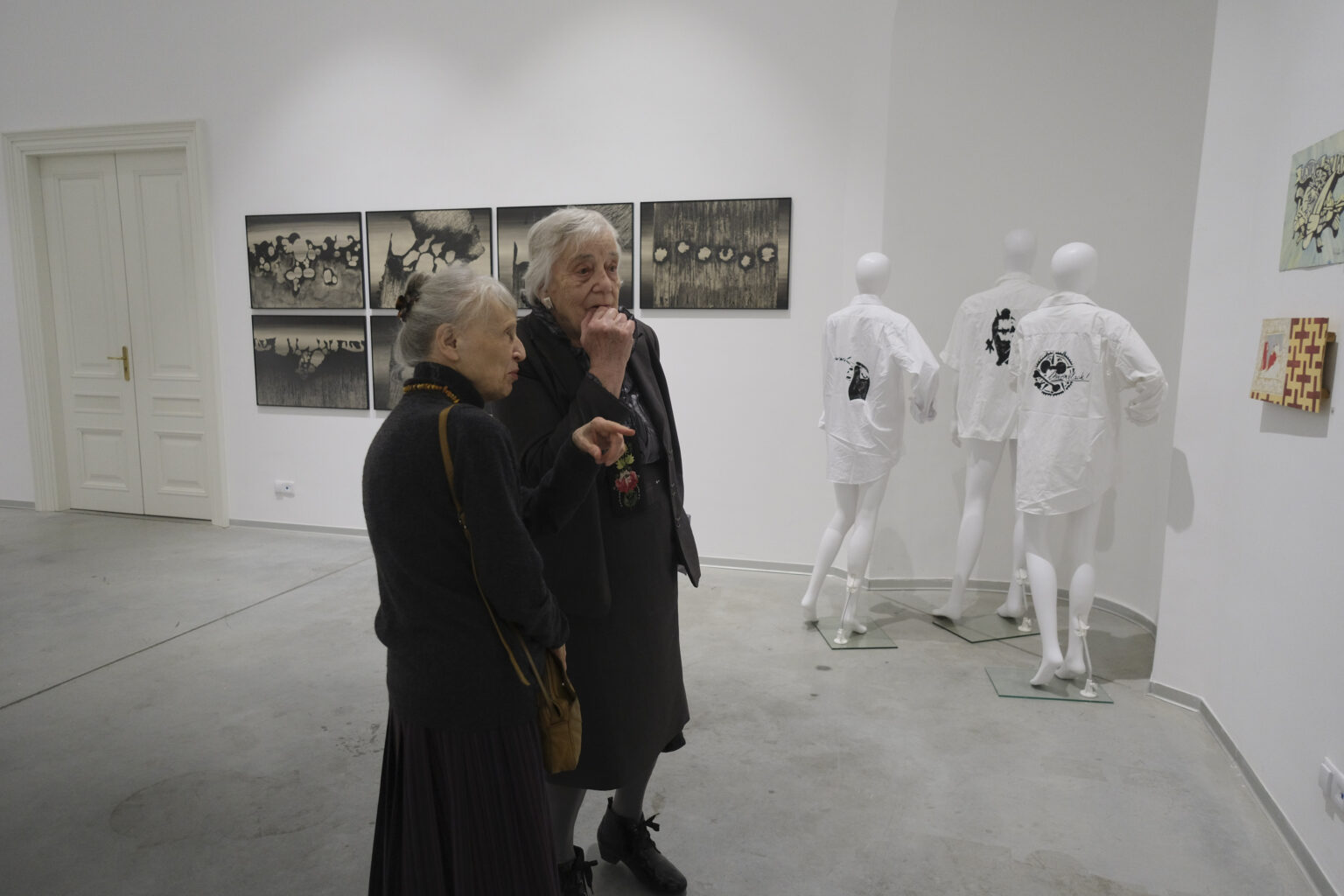 Dwie panie podziwiają prace zawieszone na ścianach w przestrzeni galerii.