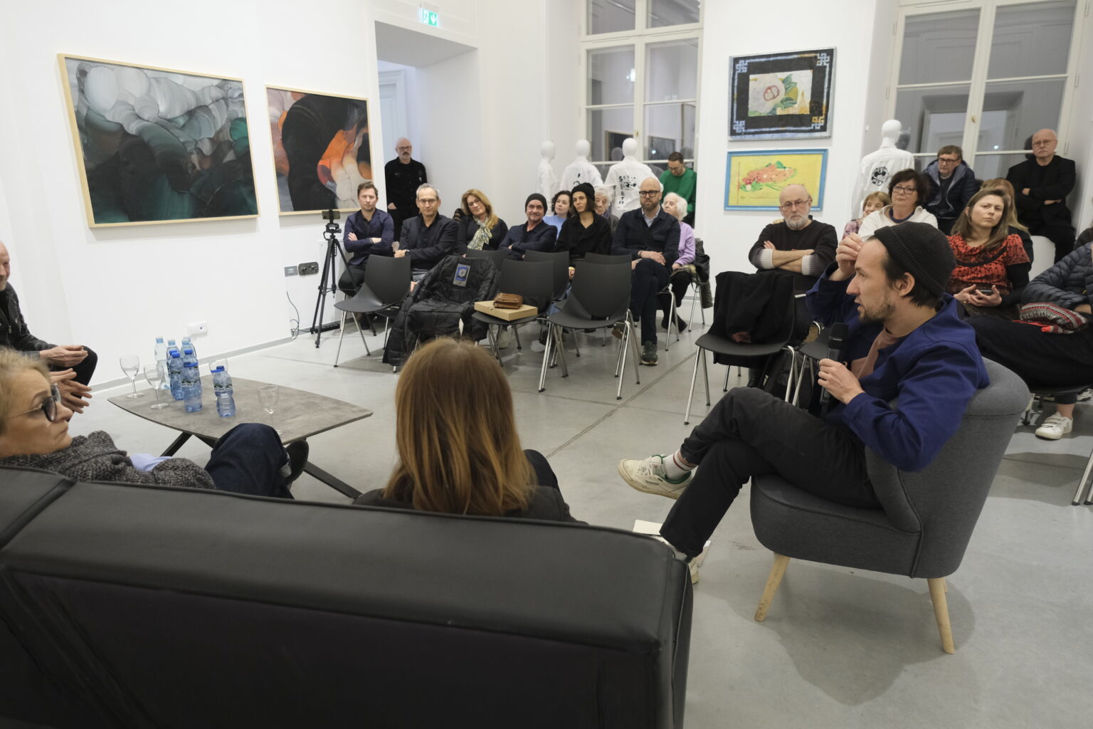 W sali z obrazami Konrada i Pawła Jarodzkich na krzesłach siedzą słuchacze debaty. Na pierwszym planie prowadzący i prelegenci.