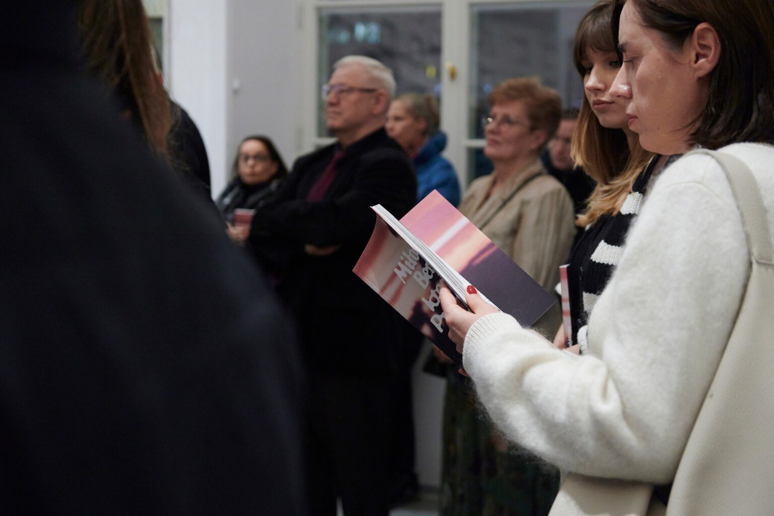 duża grupa osób w przestrzeni wystawy, na pierwszym planie osoba w białym swetrze trzymająca w dłoniach kolorowy katalog