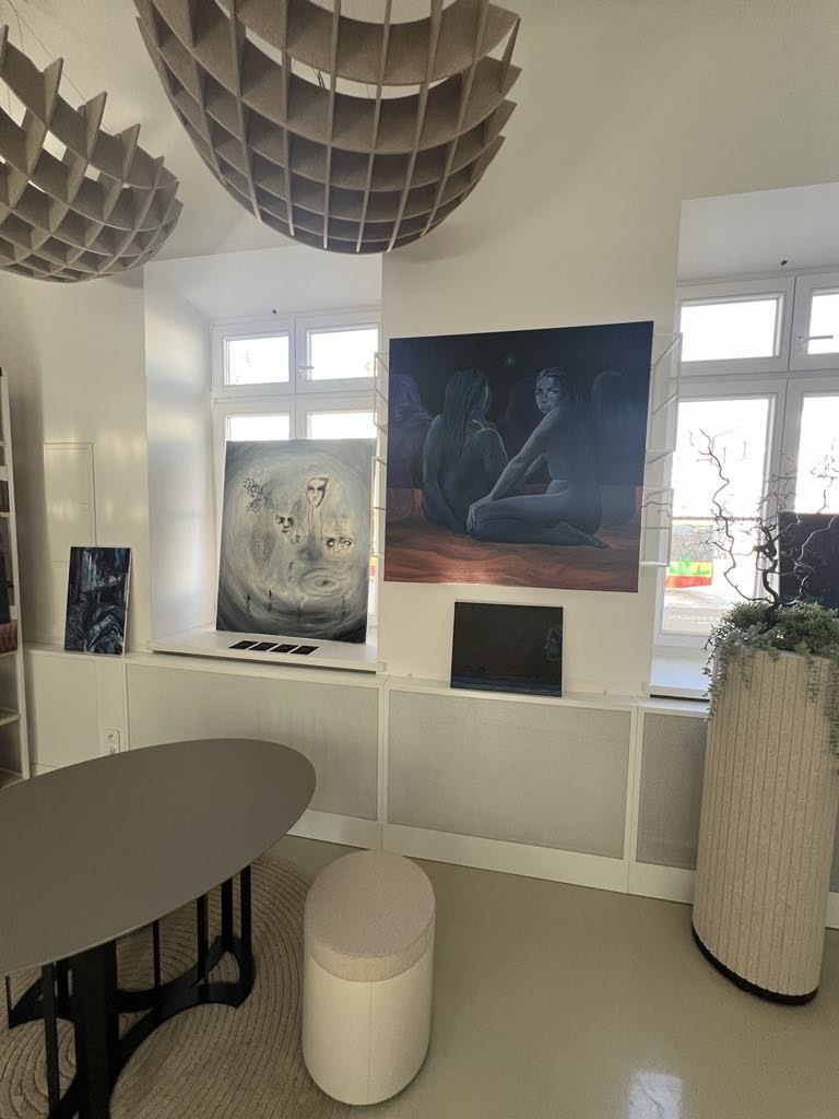 na parapecie dużego okna stoi duży obraz w kolorach bieli przedstawiający ludzkie twarze, na ścianie obok widać obraz w kolorach granatu, fioletu oraz bordo przedstawiający postacie aniołów