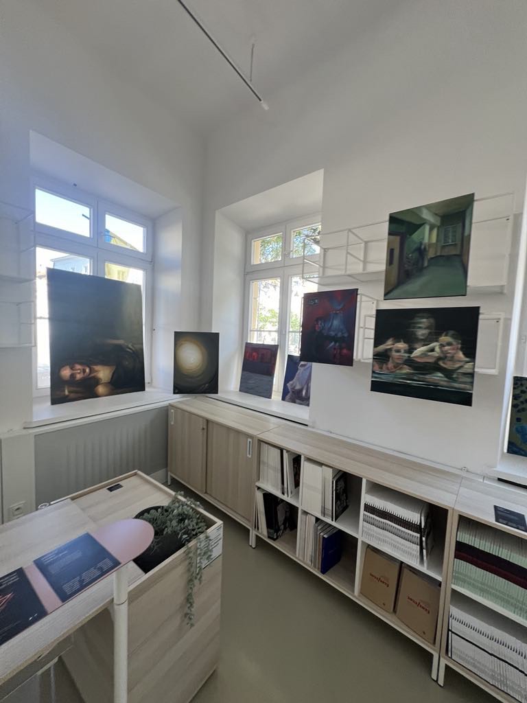 Na parapecie dużego okna stoi duży obraz w kolorach bieli przedstawiający ludzkie twarze, na ścianie obok widać obraz w kolorach granatu, fioletu oraz bordo przedstawiający postacie aniołów