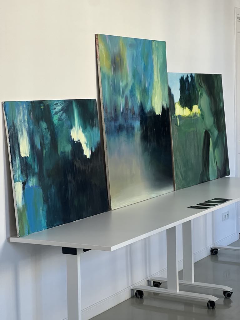 W przestrzeni wystawowej, na białych stołach, stoją trzy duże obrazy abstrakcyjne w barwach, zielni, żółci i czerni