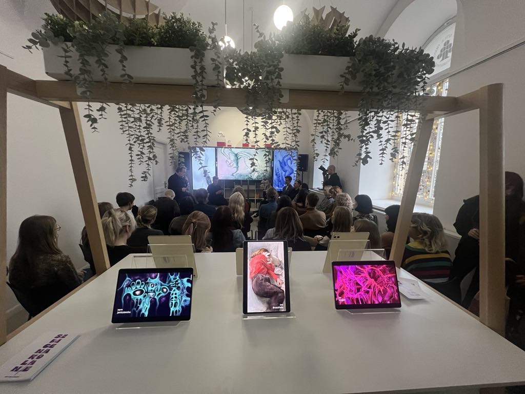 Ustawione na stole 3 laptopy, wyświetlają kolorowe prace graficzne, w oddali siedzą ludzie