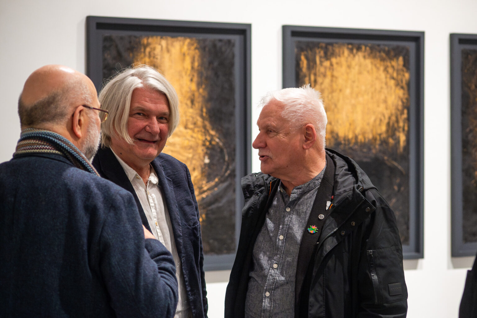 trzej mężczyźni rozmawiają na tle duzych obrazów w złotych barwach