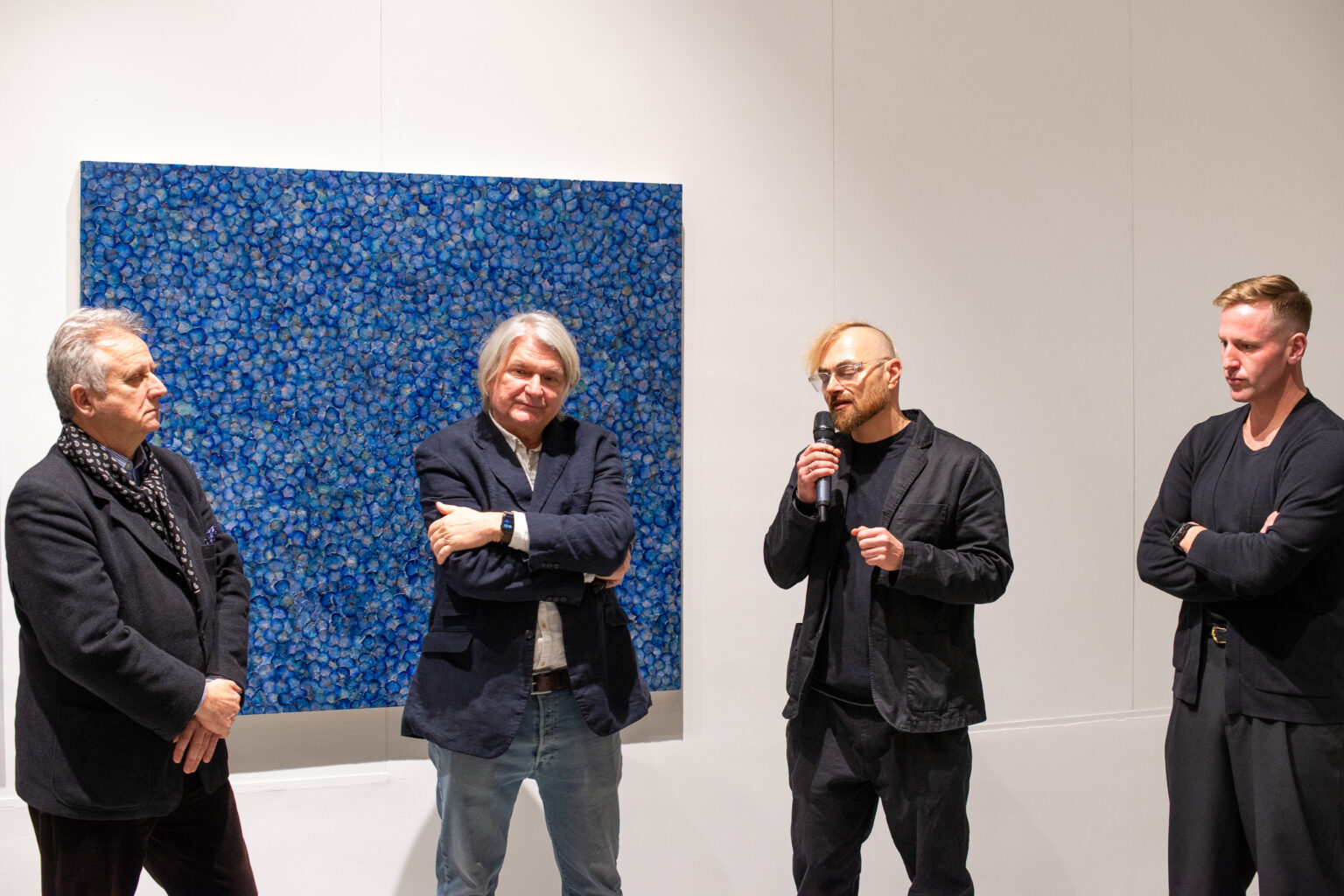 czterech mężczyzn, jeden mówi do mikrofonu, w tle dużych rozmiarów obraz abstrakcyjny