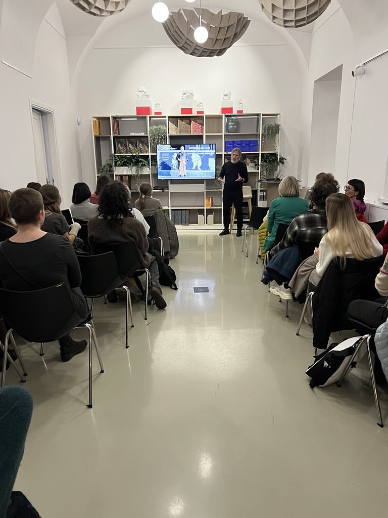 Publiczność patrzy na mężczyznę, ubranego na czarno, stoi na tle regału z książkami