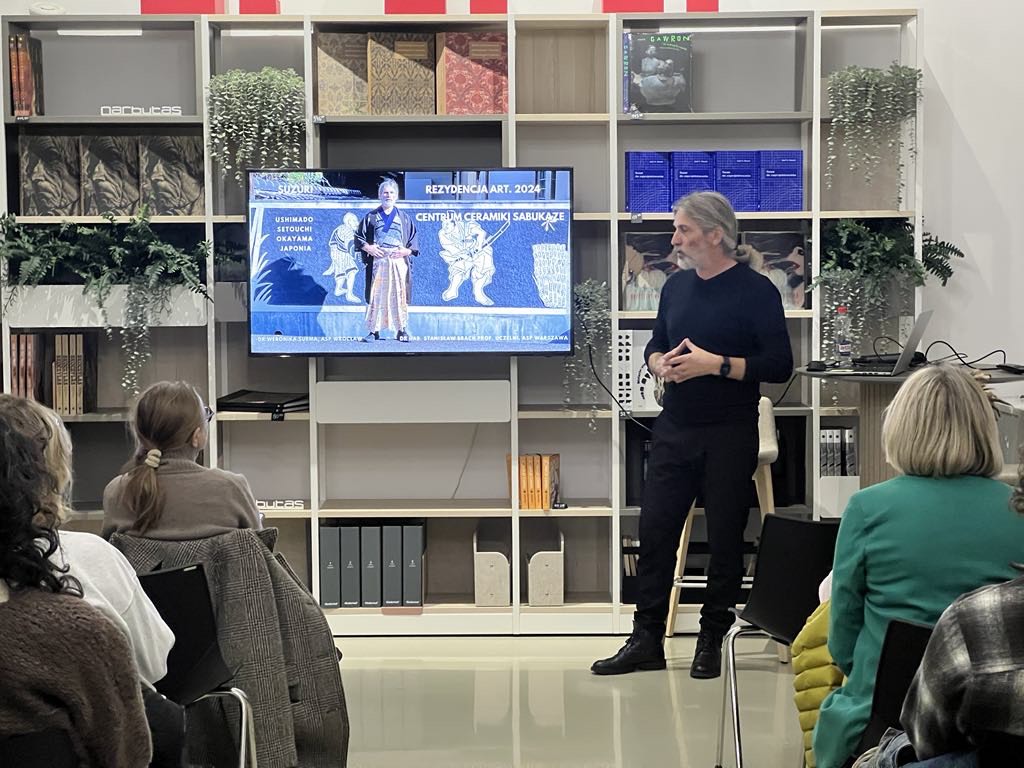 Mężczyzna ubrany na czarno, patrzy na ekran na którym widać postać ubraną w kimono.