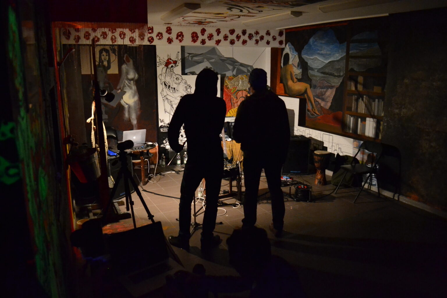 zdjęcie dwóch tyłem stojących postaci na tle sciany wypełnionej różnymi, barwnymi obrazami.