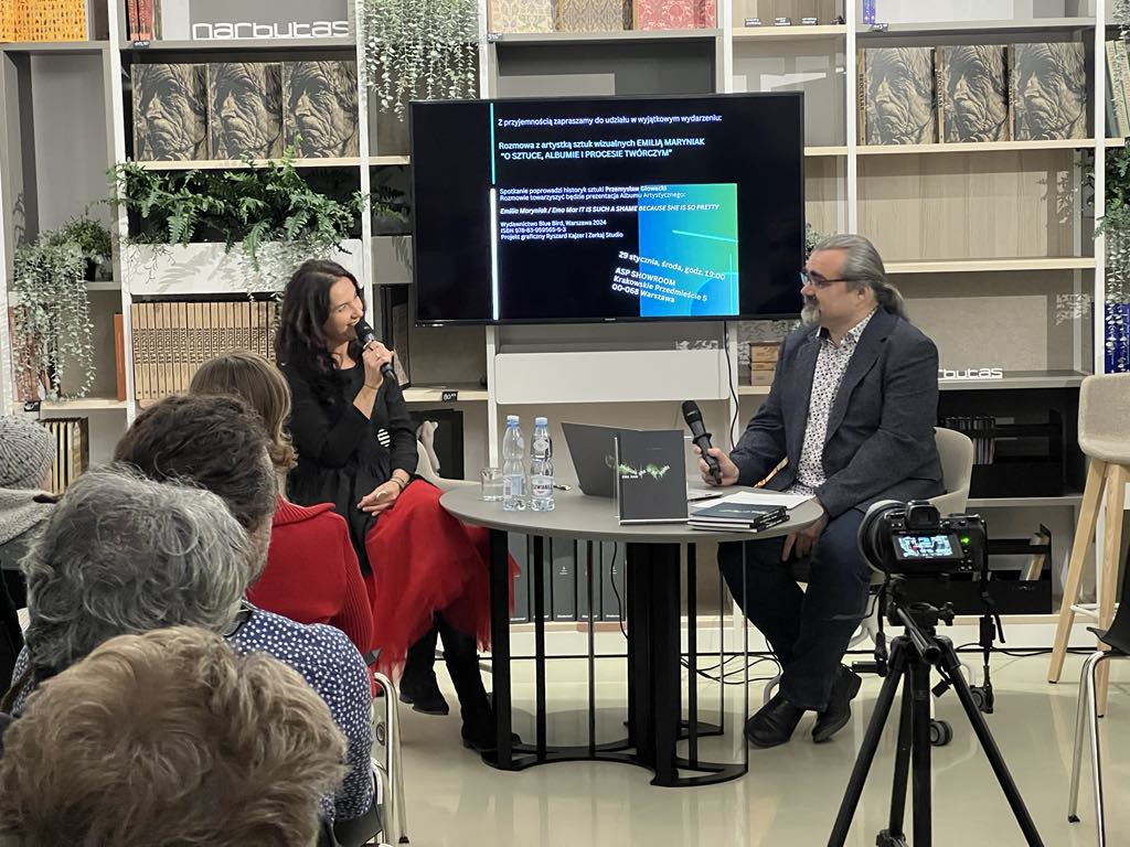 Artystka Emilia Maryniak (po lewej, na scenie) i prelegent rozmawiają podczas spotkania. Siedzą przy małym stoliku przed ekranem, a przed nimi widownia.