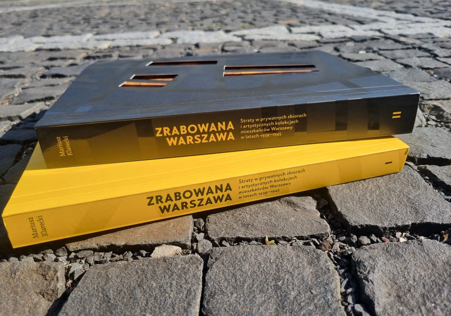 Dwa egzemplarze książki "Zrabowana Warszawa" (żółty i czarny) leżą na brukowanej ulicy w słoneczny dzień
