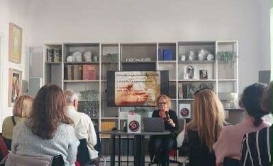 Wykład w sali z publicznością. Prelegentka siedzi za stołem, przed ekranem z prezentacją. W tle regał z książkami i rzeźbami. zdj.Agata Rachwał