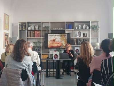 Wykład w sali z publicznością. Prelegentka siedzi za stołem, przed ekranem z prezentacją. W tle regał z książkami i rzeźbami. zdj.Agata Rachwał