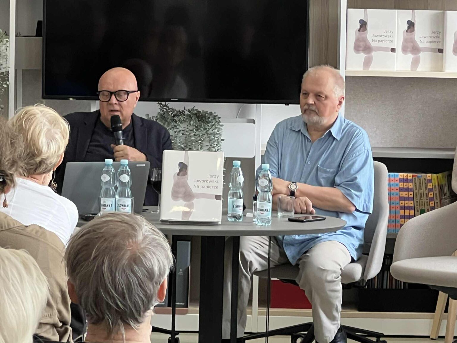 Spotkanie autorskie: dwóch mężczyzn siedzi przy stoliku, jeden z mikrofonem. Na stoliku książka "Jerzy Jaworowski. Na papierze". Publiczność. zdj.Agata Rachwał