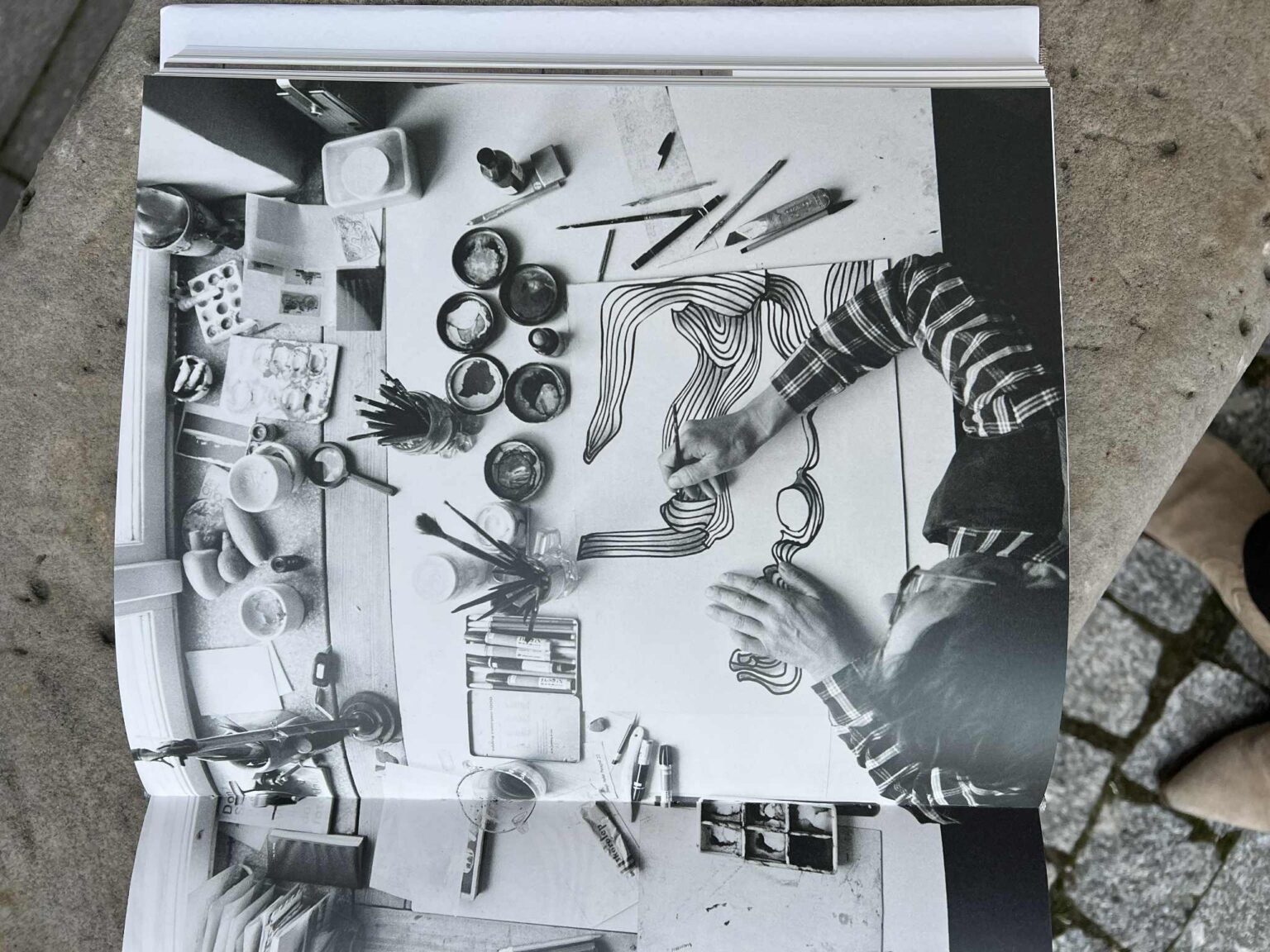 Czarno-białe zdjęcie w książce. Artysta, widziany z góry, rysuje faliste linie na kartce, otoczony farbami, pędzlami i innymi akcesoriami. zdj.Agata Rachwał