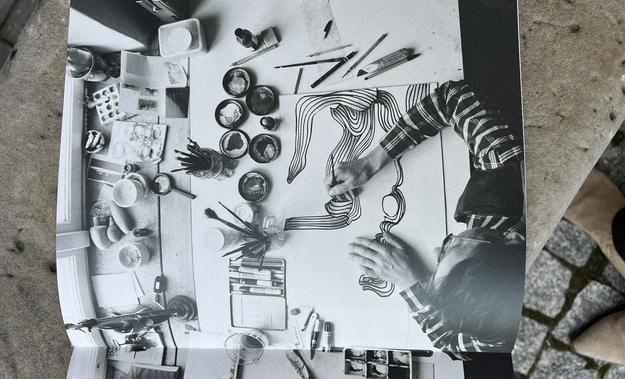 Czarno-białe zdjęcie w książce. Artysta, widziany z góry, rysuje faliste linie na kartce, otoczony farbami, pędzlami i innymi akcesoriami. zdj.Agata Rachwał