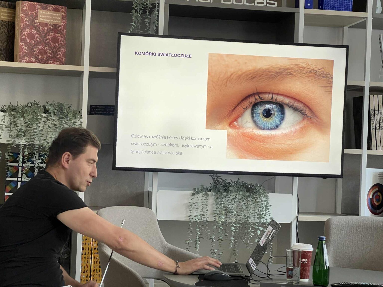 Mężczyzna przy laptopie podczas prezentacji. Na ekranie slajd "Komórki światłoczułe" i zbliżenie na niebieskie oko. W tle regały z książkami. zdj.Agata Rachwał