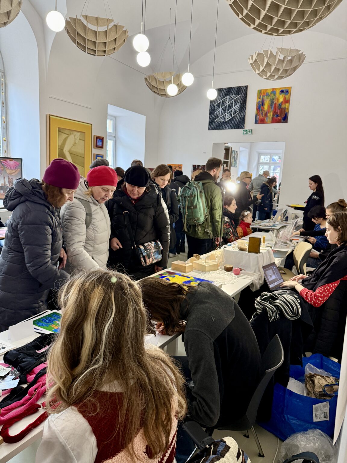Kobiety w zimowych kurtkach oglądają rękodzieło na stoisku artystycznym. W tle tłum odwiedzających, obrazy na ścianach i designerskie lampy.