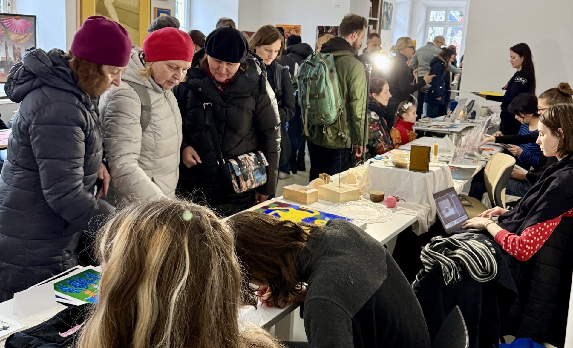 Kobiety w zimowych kurtkach oglądają rękodzieło na stoisku artystycznym. W tle tłum odwiedzających, obrazy na ścianach i designerskie lampy.