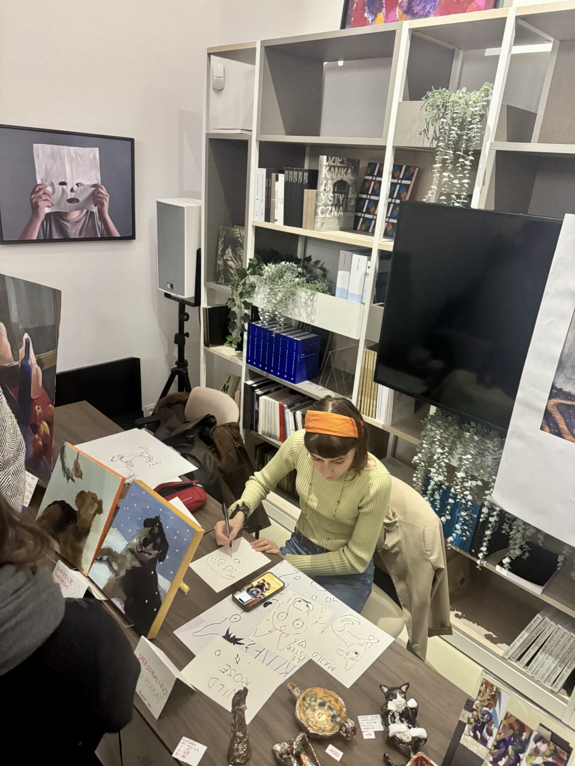 Młoda artystka w opasce rysuje przy biurku pełnym szkiców i obrazów z psami. Obok stoją ceramiczne figurki, a w tle widać regał z albumami o sztuce.