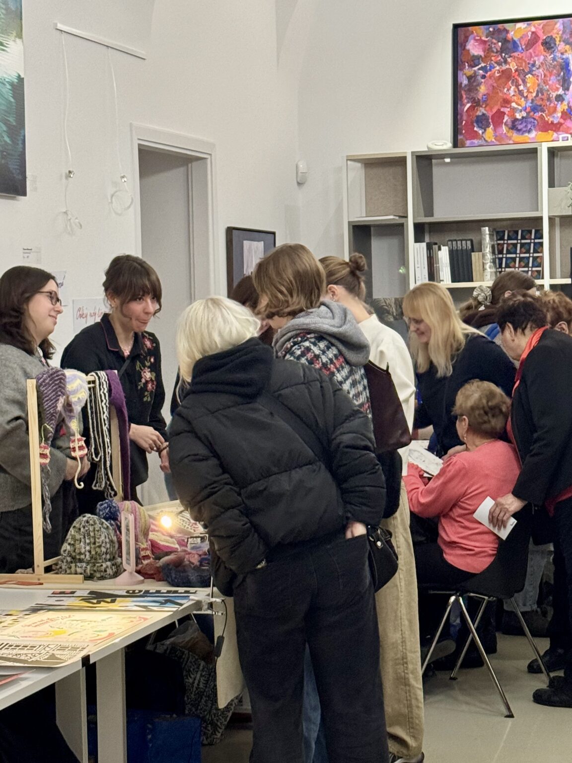 Artystki rozmawiają z klientkami przy stoisku z dzianinami i biżuterią. W tle widać kolorowe obrazy na ścianach oraz innych odwiedzających targi sztuki.