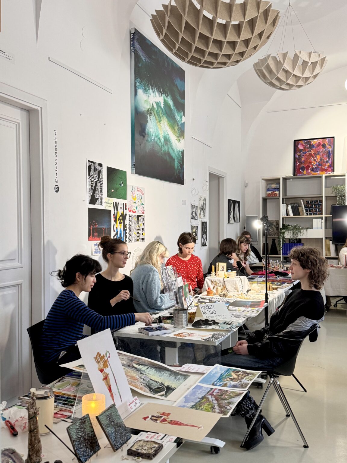 Młodzi ludzie przy stołach pełnych grafik i obrazów w jasnym, wysokim wnętrzu. Na pierwszym planie artystka maluje portret osoby pozującej na krześle.