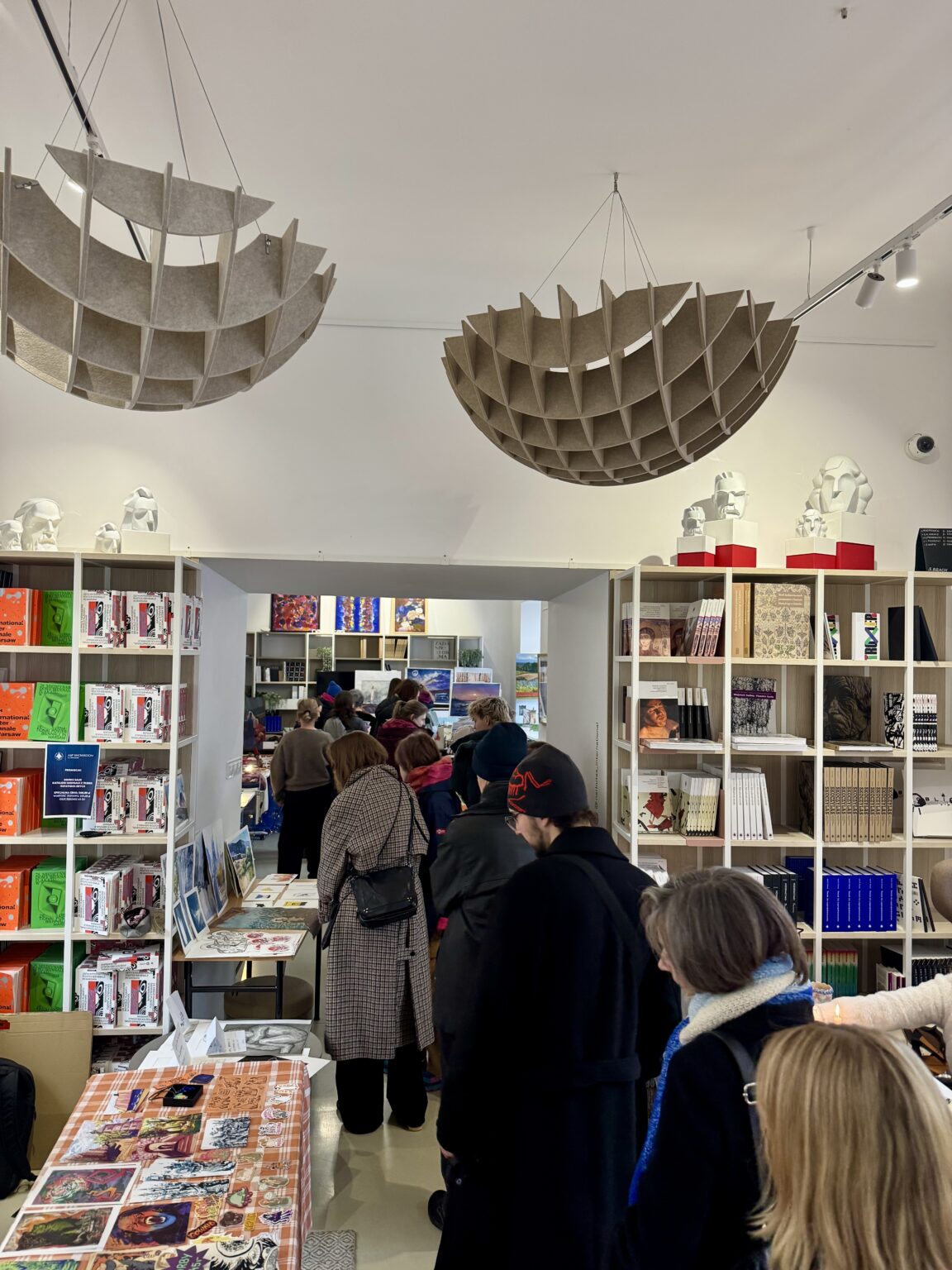 Tłum gości przechodzi między regałami pełnymi książek i grafik. Nad przejściem wiszą duże, ażurowe lampy, a na szczycie mebli stoją białe popiersia.