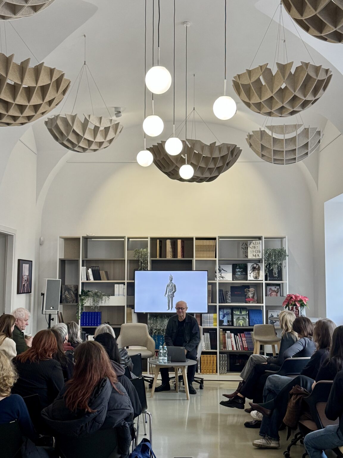 Na tle dużej półki z książkami stoi Maciej Buszewicz, przemawiający do widowni. zdj.Anita Lewandowska