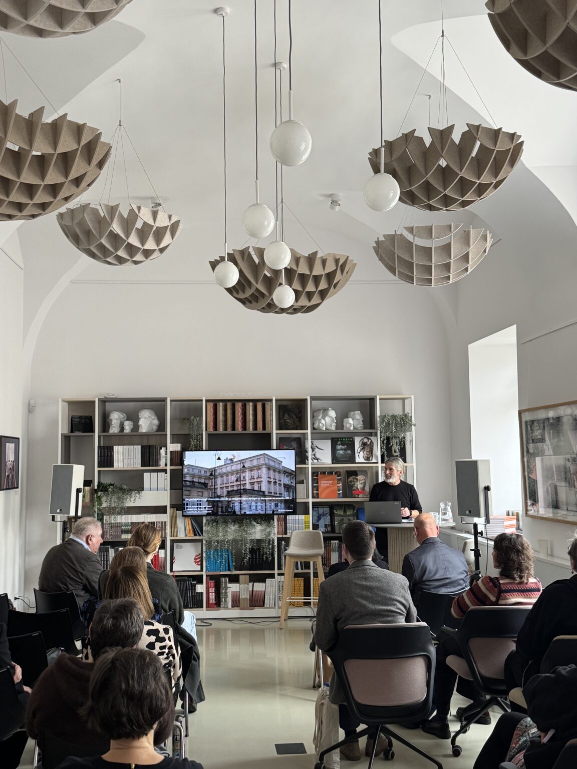 Wysokie, jasne pomieszczenie, na tle książek i rzeźb widnieje ekran pokazujący budynek obok z laptopem stoi prowadzący. zdj. Anita Lewandowska