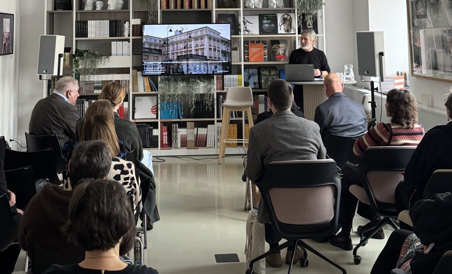 Wysokie, jasne pomieszczenie, na tle książek i rzeźb widnieje ekran pokazujący budynek obok z laptopem stoi prowadzący. zdj. Anita Lewandowska